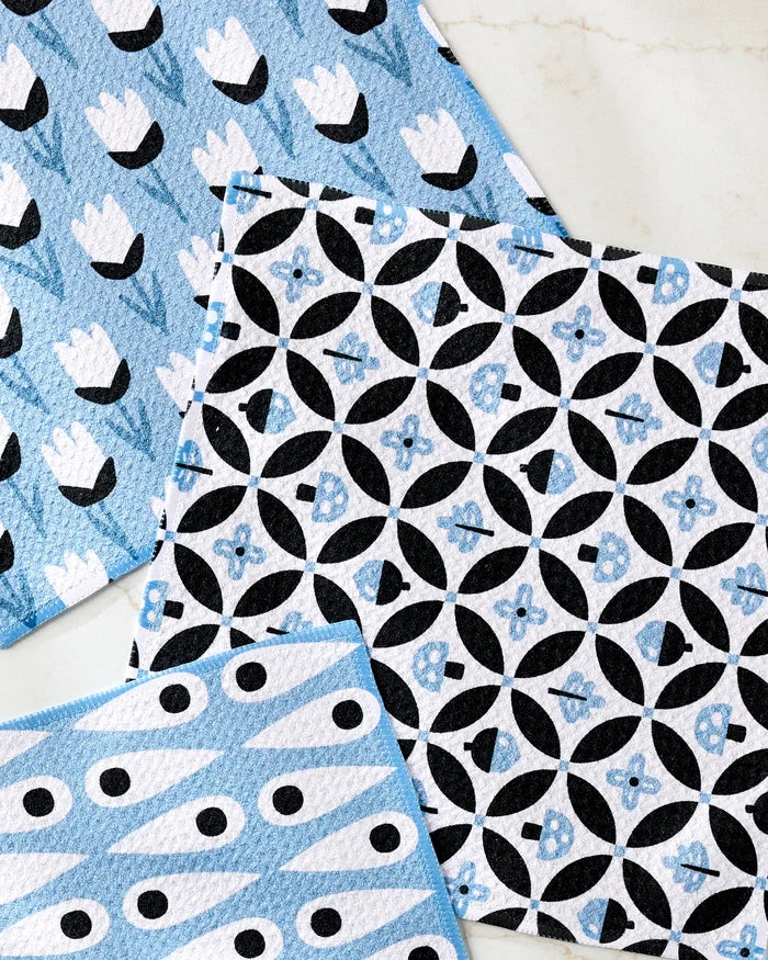 Three "Forest Findings" dishcloths by Anette Heiberg, made from recycled materials, display blue, black, and white floral, circular, and abstract eye designs on a light marble surface.