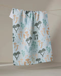 The "Forest Friends" kitchen towel by Amberleigh Adoff features a whimsical mushroom pattern in green, orange, and beige on light blue, displayed hanging on a clothesline against a neutral background.