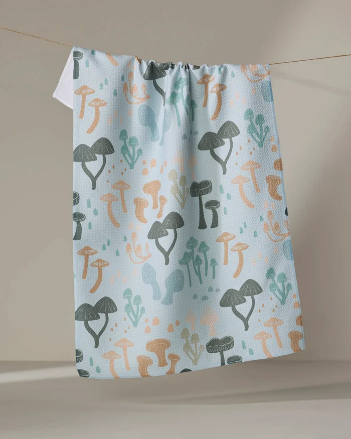 The "Forest Friends" kitchen towel by Amberleigh Adoff features a whimsical mushroom pattern in green, orange, and beige on light blue, displayed hanging on a clothesline against a neutral background.
