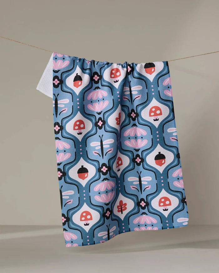 The Anette Heiberg "Forest Garden" dish towel, featuring a blue, pink, and red abstract floral and mushroom pattern, hangs on a line against a neutral background.