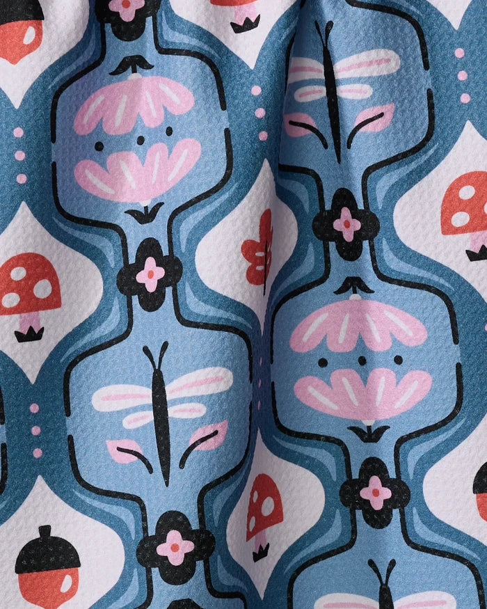 Forest Garden by Anette Heiberg is a colorful recycled kitchen towel with a blue background, featuring black, pink, red, and white dragonflies, mushrooms, acorns, and stylized flowers. The fabric has a slightly wrinkled look.