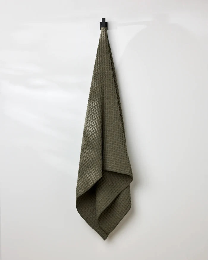 The GEOMETRY Forest bath towel in olive green, featuring a textured waffle pattern, hangs neatly on a black hook against a plain white wall, with soft folds and gentle shadows accentuating its design.
