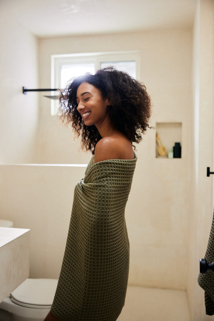 A woman with curly hair smiles in a sunlit bathroom, wrapped in the GEOMETRY Forest textured green waffle bath towel, with eco-friendly Forest towels stacked neatly on a shelf behind her.