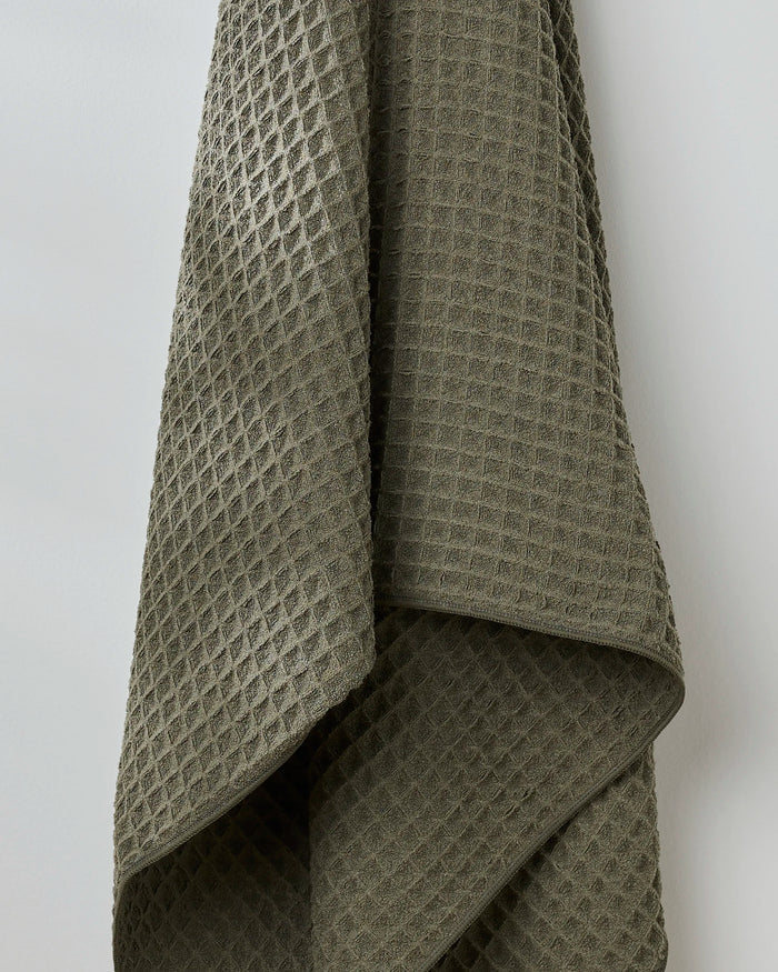 The GEOMETRY Forest towel, featuring a textured olive-green waffle weave, hangs against a plain white wall and brings understated luxury to your space.
