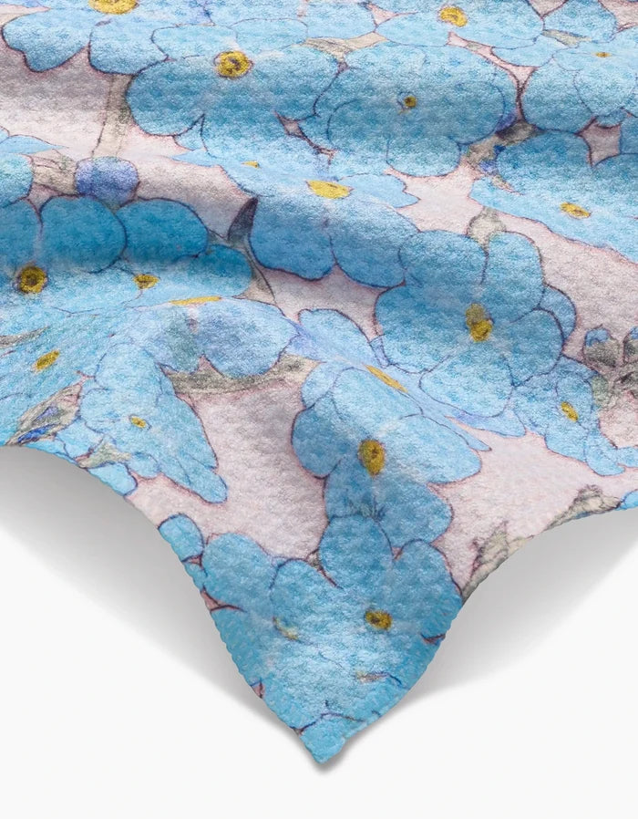 A close-up of Pascale Dilgers Forget Me Not Floral Bar Towel shows its textured Geoweave fabric with blue flowers and yellow centers on a light background, and a wavy edge visible at the bottom.