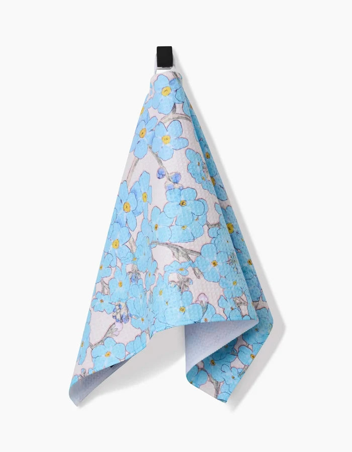 The Pascale Dilger Forget Me Not Floral bar towel, featuring a blue floral pattern with yellow accents and crafted from Geoweave fabric, hangs from a black loop against a plain white background.