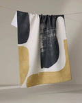 The Geometry Forma dish towel, featuring quick-drying fabric and abstract black, white, and gold shapes, hangs draped over a clothesline against a plain light background.