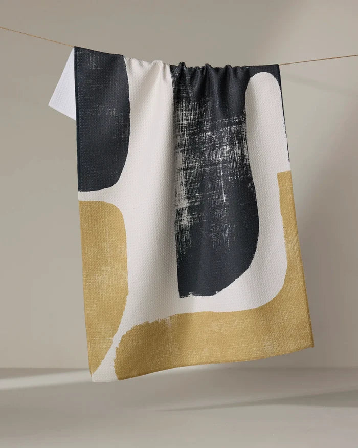 The Geometry Forma dish towel, featuring quick-drying fabric and abstract black, white, and gold shapes, hangs draped over a clothesline against a plain light background.