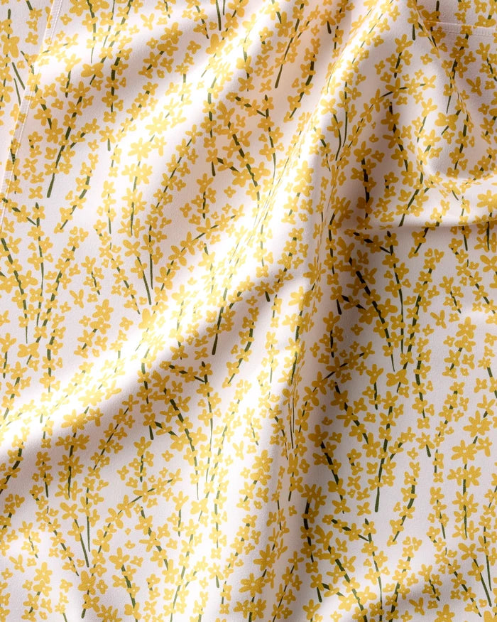 A close-up of Kayla Ann’s Forsythia Floral eco-friendly table linens shows yellow branches and leaves on a white background, with the softly wrinkled fabric creating gentle folds and shadows.