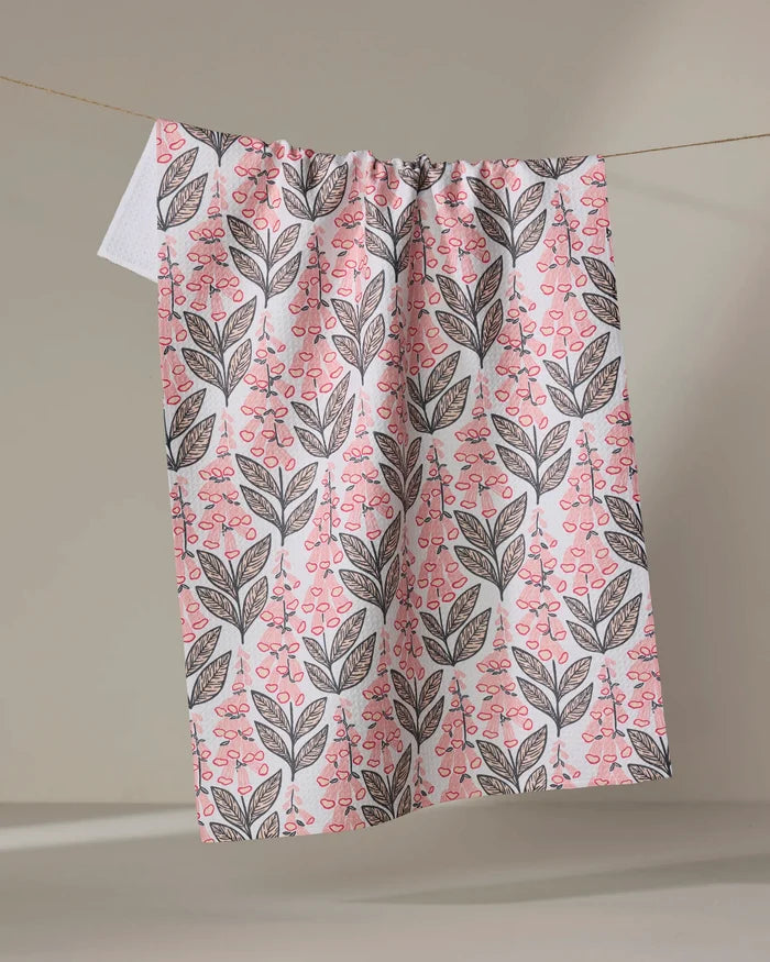 The Foxglove Finery kitchen towel by Sarah Daugherty features a pink and black botanical leaf and berry pattern on white eco-friendly fabric, hanging on a clothesline with a neutral backdrop.