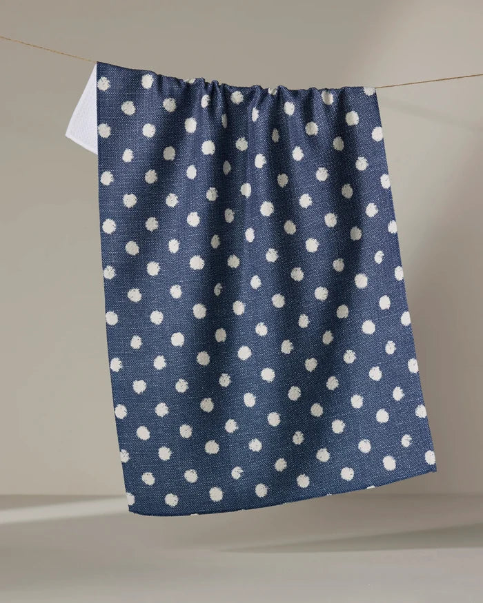 The Geometry Freckle Jean, a navy blue tea towel with white polka dots made from recycled materials, hangs over a thin clothesline against a plain beige background.