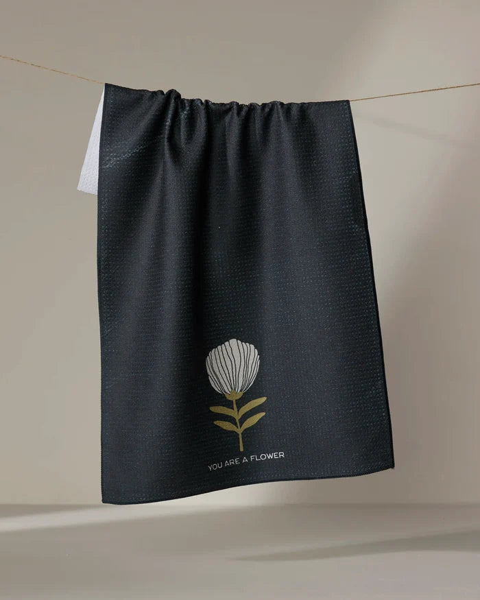 The Freja tea towel by Anna Christian is made from recycled materials, featuring a simple white and yellow flower illustration with the text "YOU ARE A FLOWER," shown hanging against a neutral background.