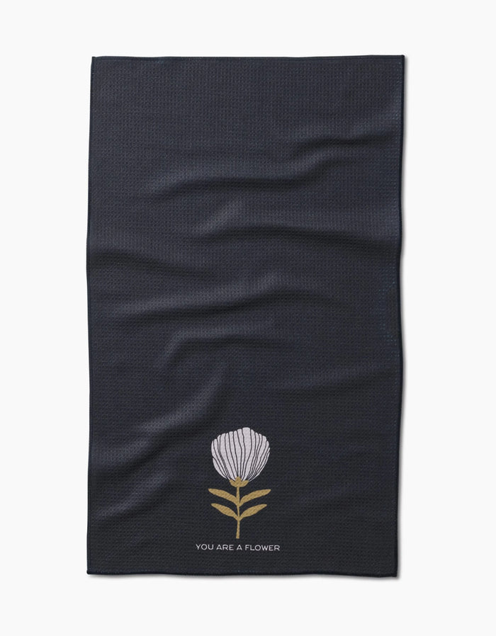 The Freja tea towel by Anna Christian is made from dark Geoweave fabric, featuring a single white flower with green leaves and YOU ARE A FLOWER printed below the illustration.
