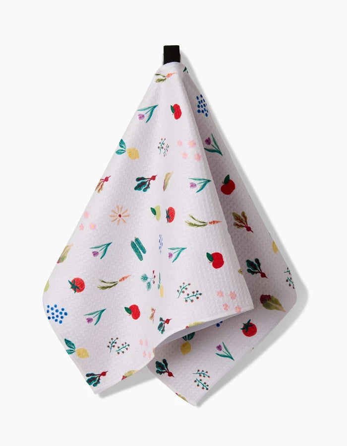 The Fresh Produce towel by Brittany Jepsen, made from ultra-absorbent Geoweave fabric, features a colorful fruit, leaf, and flower pattern on white and hangs from a black loop against a plain background.
