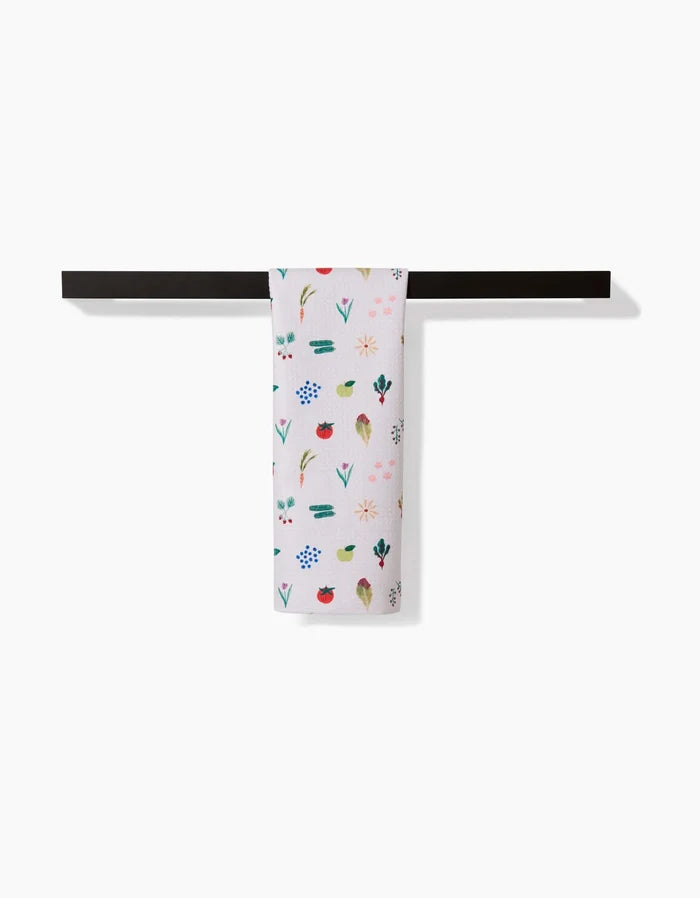 The Brittany Jepsen Fresh Produce dish towel, made from ultra-absorbent Geoweave fabric and featuring colorful floral and leaf patterns, hangs neatly on a black rod against a plain white background.