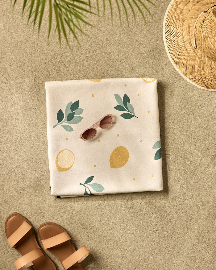 The Sara Mari Freshly Picked micro-loop beach blanket featuring a lemon and leaf pattern is spread on the sand with pink sunglasses, a straw hat, and brown sandals beneath the shadow of a palm leaf.