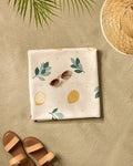 The Sara Mari Freshly Picked micro-loop beach blanket featuring a lemon and leaf pattern is spread on the sand with pink sunglasses, a straw hat, and brown sandals beneath the shadow of a palm leaf.