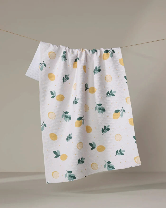 The "Freshly Picked" tea towel by Sara Mari, featuring a yellow lemon and green leaf pattern on white textured recycled fabric, hangs on a string against a light background.
