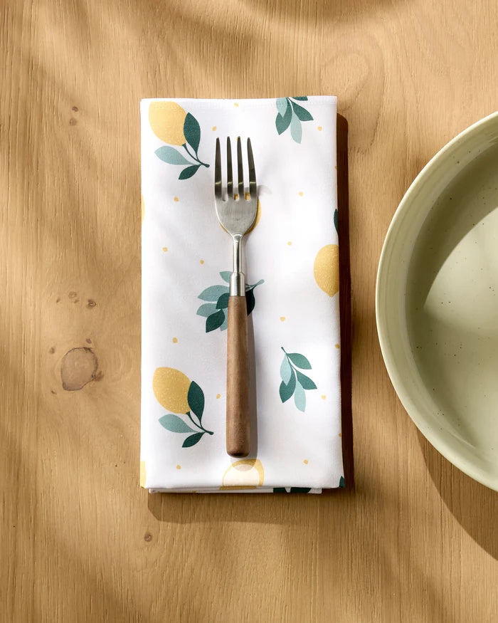 A Sara Mari "Freshly Picked" lemon-and-leaf dinner napkin, topped with a wooden-handled fork, lies by a bowl on a wooden table. Sunlight adds gentle shadows to the bright, cheerful scene.
