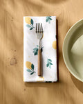 A Sara Mari "Freshly Picked" lemon-and-leaf dinner napkin, topped with a wooden-handled fork, lies by a bowl on a wooden table. Sunlight adds gentle shadows to the bright, cheerful scene.