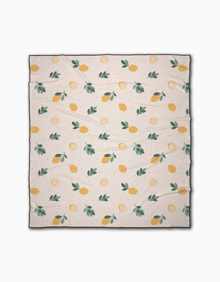 The Sara Mari Freshly Picked beach blanket is cream-colored, quick-drying, and made from recycled materials. It features a repeating pattern of yellow lemons, lemon slices, and green leaves with slight visible wrinkles when spread out.