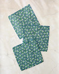 Three green Friendly Fleurs fabric napkins by Megan Kampa, featuring a white, blue, and yellow floral pattern, are displayed on a light beige marble surface; they’re crafted from recycled materials for eco-friendly style.