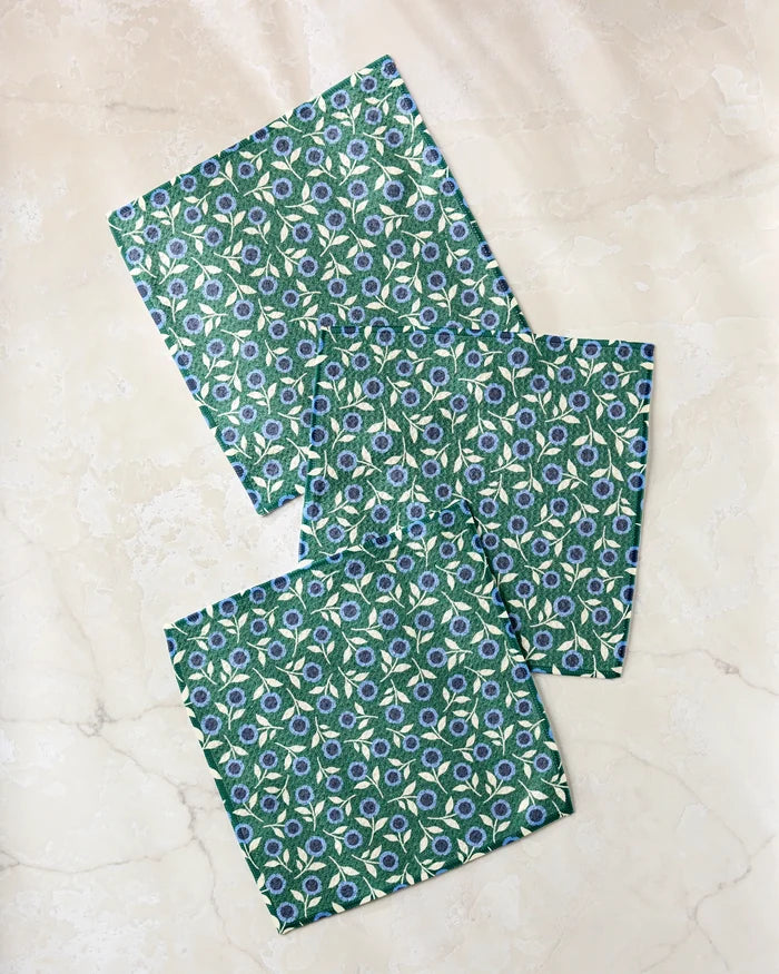 Three green Friendly Fleurs fabric napkins by Megan Kampa, featuring a white, blue, and yellow floral pattern, are displayed on a light beige marble surface; they’re crafted from recycled materials for eco-friendly style.