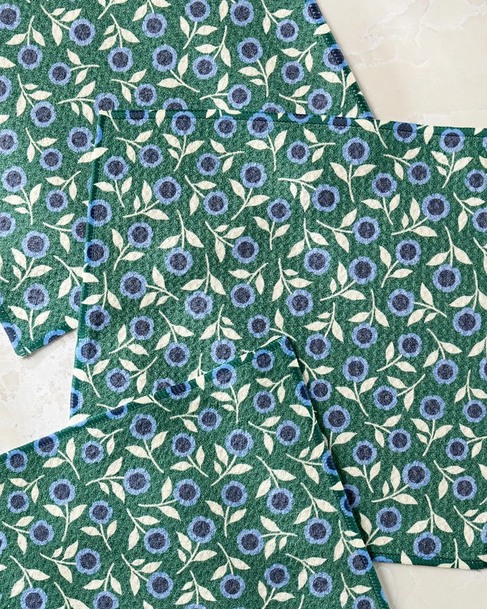 Four Friendly Fleurs cloths by Megan Kampa, made from recycled materials, are displayed on a light surface. Each green cloth features a repeating pattern of blue and white flowers with white stems and leaves.
