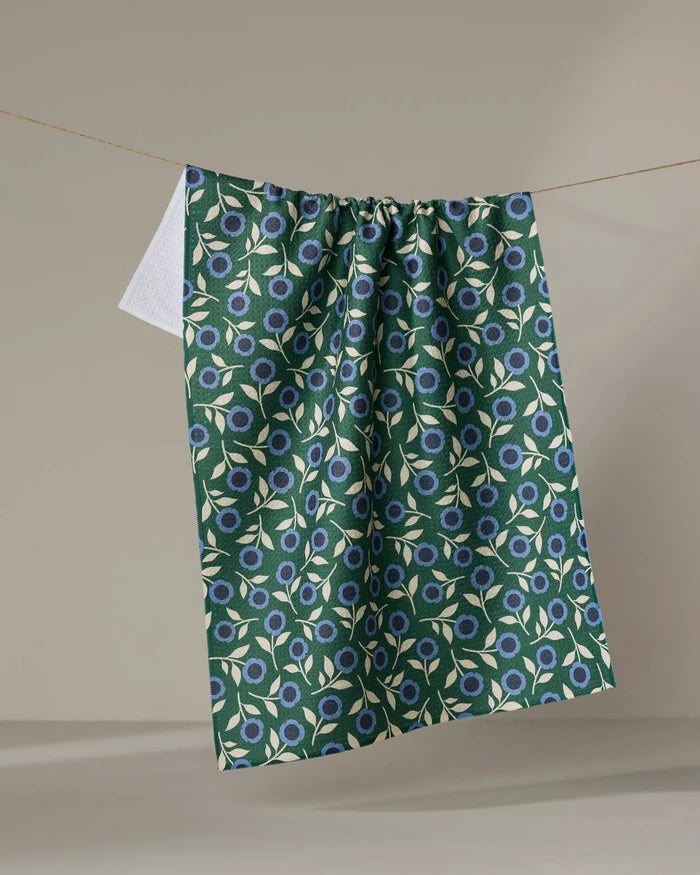 The Megan Kampa 'Friendly Floral Blue' tea towel, made from recycled materials and featuring blue flowers with white leaves on green fabric, hangs on a line against a plain, neutral background.