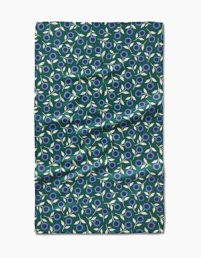 The Megan Kampa Friendly Floral Blue tea towel is rectangular, made from recycled materials, and features a dark green background with blue flowers and white leaves. Slightly wrinkled fabric is photographed on a white surface.