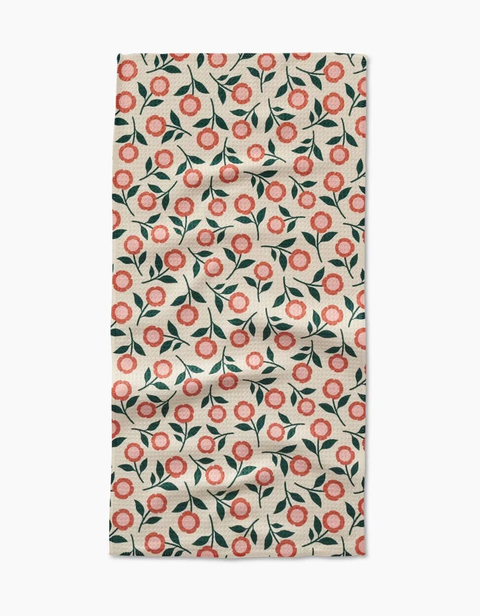 A slightly wrinkled Megan Kampa Friendly Floral Peach Geoweave bar towel, with a repeating red and pink flower and green leaf pattern on cream, is displayed on a white surface.
