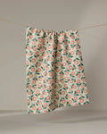 The "Friendly Floral Peach" tea towel by Megan Kampa, made from recycled materials and adorned with pink and green flowers, hangs from a clothesline against a neutral background.