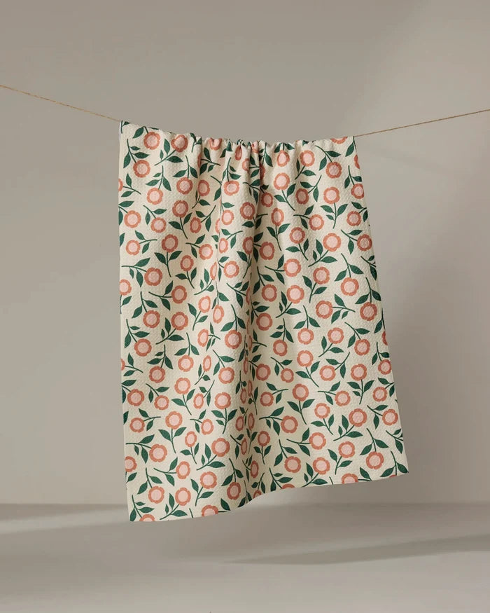The "Friendly Floral Peach" tea towel by Megan Kampa, made from recycled materials and adorned with pink and green flowers, hangs from a clothesline against a neutral background.