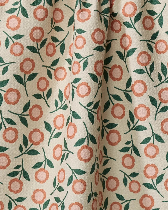 A close-up of Megan Kampa's Friendly Floral Peach tea towel, made from recycled materials, shows pinkish-orange flowers and green leaves on cream fabric with a textured surface and gentle folds. Ultra-absorbent and eco-friendly.