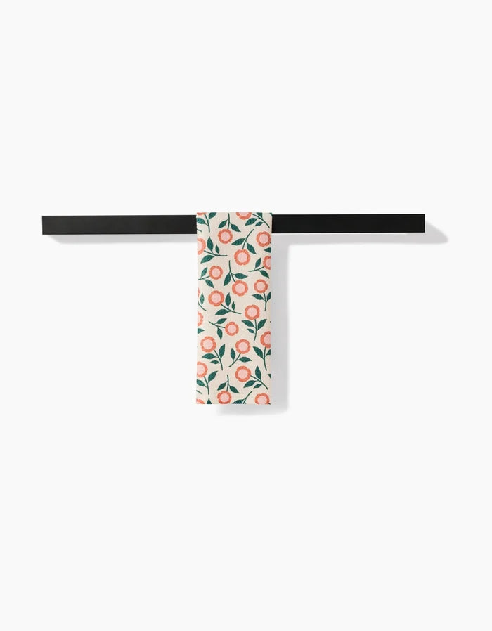 The Friendly Floral Peach bar towel by Megan Kampa, featuring a pink and green floral Geoweave fabric, hangs neatly over a minimalist black rod against a white background.