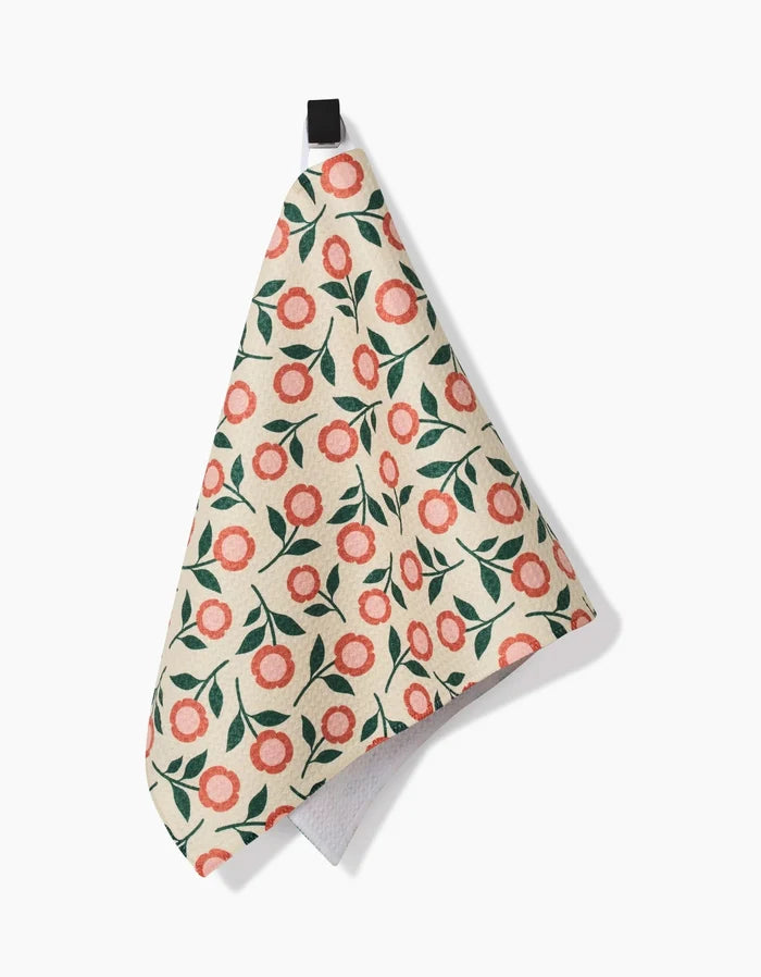The Megan Kampa Friendly Floral Peach Bar Towel, crafted from Geoweave fabric and featuring pink flowers with green leaves on a cream background, hangs by a black loop against a white backdrop.