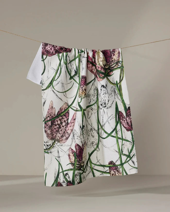 The Pascale Dilger "Fritillaria" tea towel, crafted from recycled materials, features a vibrant floral pattern of purple and white flowers with green stems and hangs on a clothesline against a plain background.
