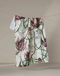 The Pascale Dilger "Fritillaria" tea towel, crafted from recycled materials, features a vibrant floral pattern of purple and white flowers with green stems and hangs on a clothesline against a plain background.