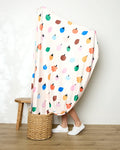 A person stands with their legs visible, wrapped in the colorful Jennifer Bouron Fruit Basket blanket adorned with fruit and flower patterns. They wear white sneakers next to a wooden stool and a woven basket on a wooden floor.