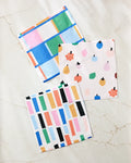 Three colorful Fruit Basket dishcloths by Jennifer Bouron rest on marble: one sports blue, green, pink, and beige checks; another has a fruit print; the third features multicolored vertical rectangles.