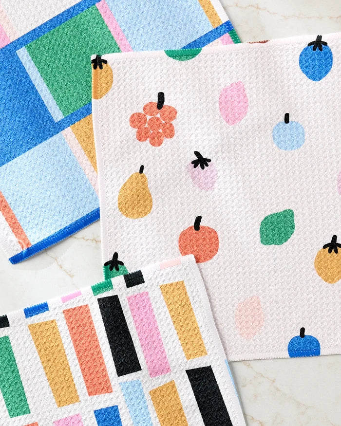 Three Fruit Basket dishcloths by Jennifer Bouron, made from recycled materials, are displayed on light marble. Each features a playful design: geometric patterns, abstract dots, and fruit illustrations in pastel and bold colors.
