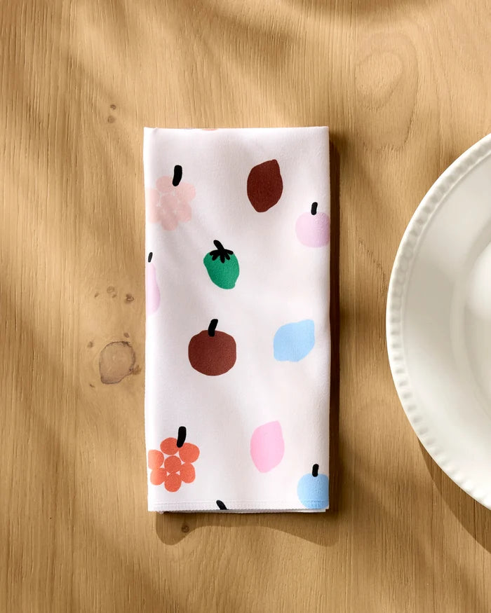 The Jennifer Bouron Fruit Basket napkin, featuring colorful abstract fruit and flower designs on white fabric, is folded on a light wooden table beside a scalloped white plate as sunlight casts gentle shadows.