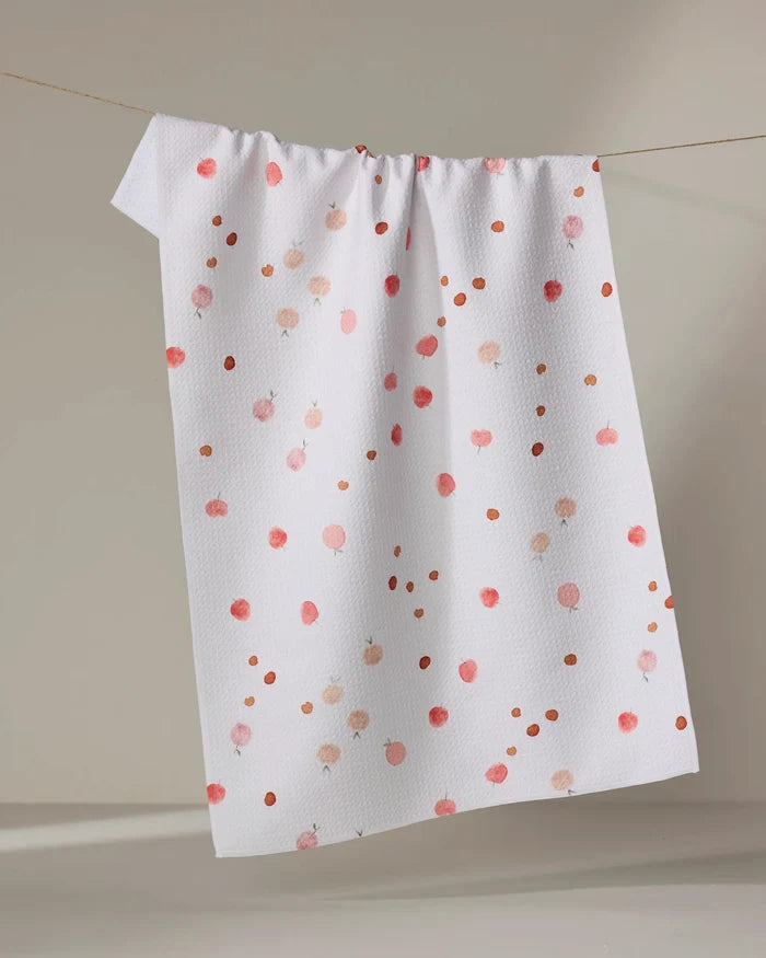 The Fruity tea towel by Anee Shah features a white textured design with pink and orange dot patterns, made from recycled materials, shown hanging on a clothesline against a neutral background.