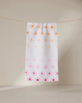 The GEOMETRY Fun in the Sun bar towel, featuring orange and pink sun patterns on white, hangs on a thin clothesline against a beige background.
