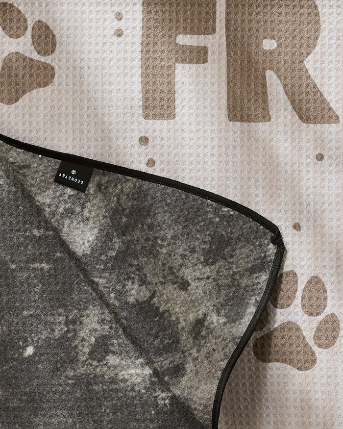 A close-up of a textured mat with brown paw prints and large "FR" letters, featuring a folded corner of the highly absorbent, marbled-gray Fur Ever Friends dog towel from Geometry, made with recycled materials.