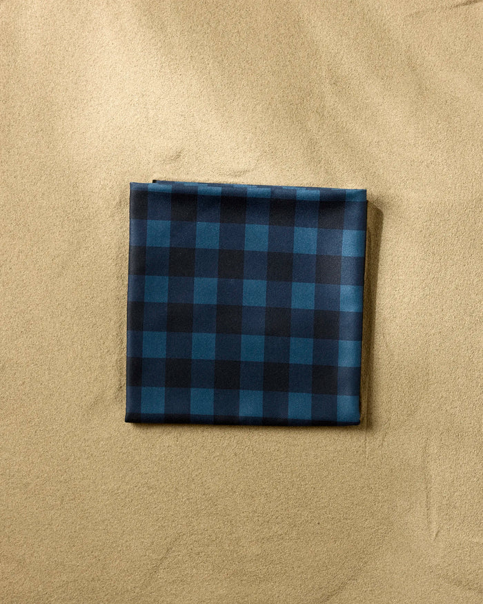 The Modern Dad’s Game Day beach blanket, made from recycled materials with a dark blue and black checkered design, lies neatly folded on smooth beige sand.