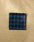 The Modern Dad’s Game Day beach blanket, made from recycled materials with a dark blue and black checkered design, lies neatly folded on smooth beige sand.