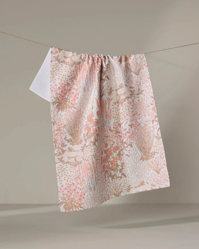 The Hufton Studio Garden Bloom tea towel, featuring a soft pink and beige floral pattern and made from recycled materials, hangs on a clothesline against a neutral backdrop.