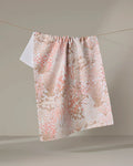 The Hufton Studio Garden Bloom tea towel, featuring a soft pink and beige floral pattern and made from recycled materials, hangs on a clothesline against a neutral backdrop.