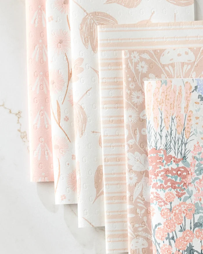 Five sheets of Hufton Studio's Garden Bloom recycled paper towels are fanned out on marble, featuring pastel floral, leaf, stripe, and mushroom patterns in pink, peach, and cream.